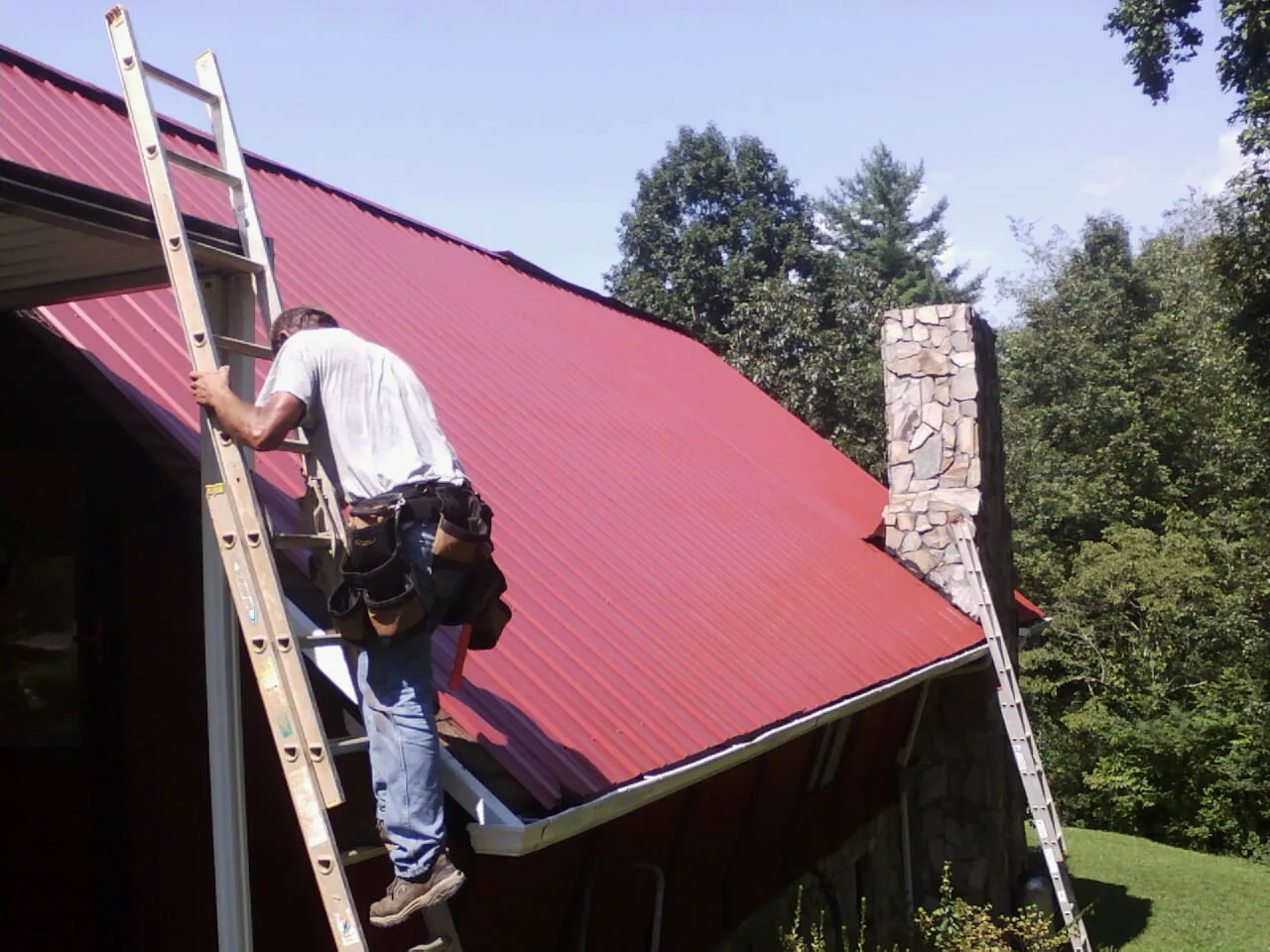 Roofer on a ladder beside a red metal roof and stone chimney — Soffit & Fascia Repair by Roofing Brothers in Selinsgrove